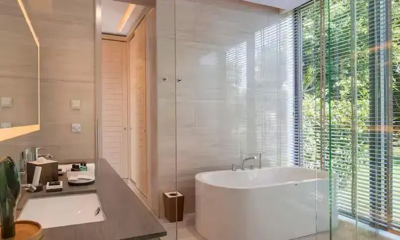 Badezimmer mit freistehender Badewanne, Glaswand und bodentiefem Fenster mit Jalousie.