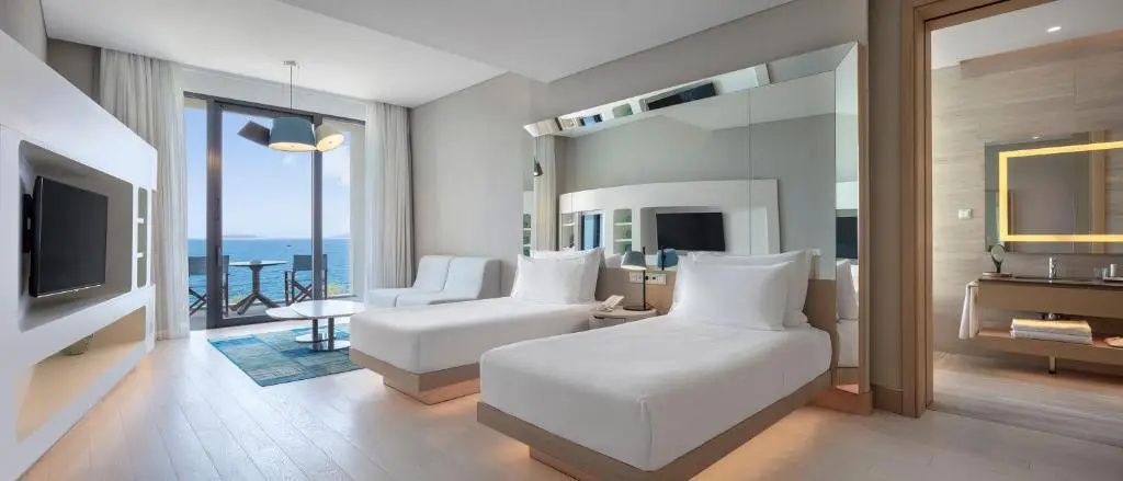 Helles Hotelzimmer mit zwei Einzelbetten, weißer Einrichtung und Blick auf das Meer