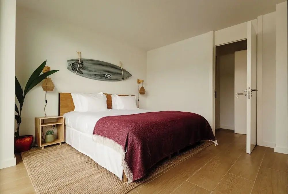 Doppelbett mit rotbrauner Tagesdecke und einem Surfbrett als Wanddekoration im hellem Zimmer.