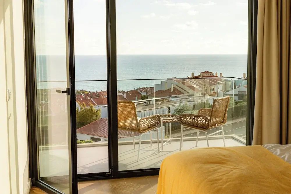 Blick von einem Balkon mit zwei Stühlen auf rote Dächer und das blaue Meer.