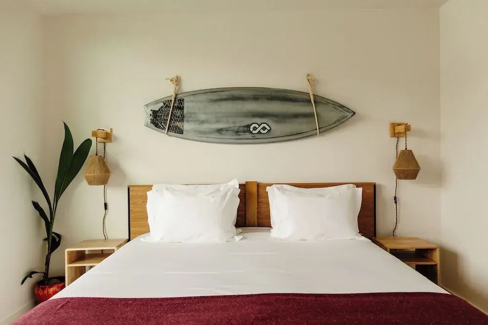 Frontalansicht des Doppelbetts mit weißer Bettwäsche und einem Surfbrett an der hellen Wand.