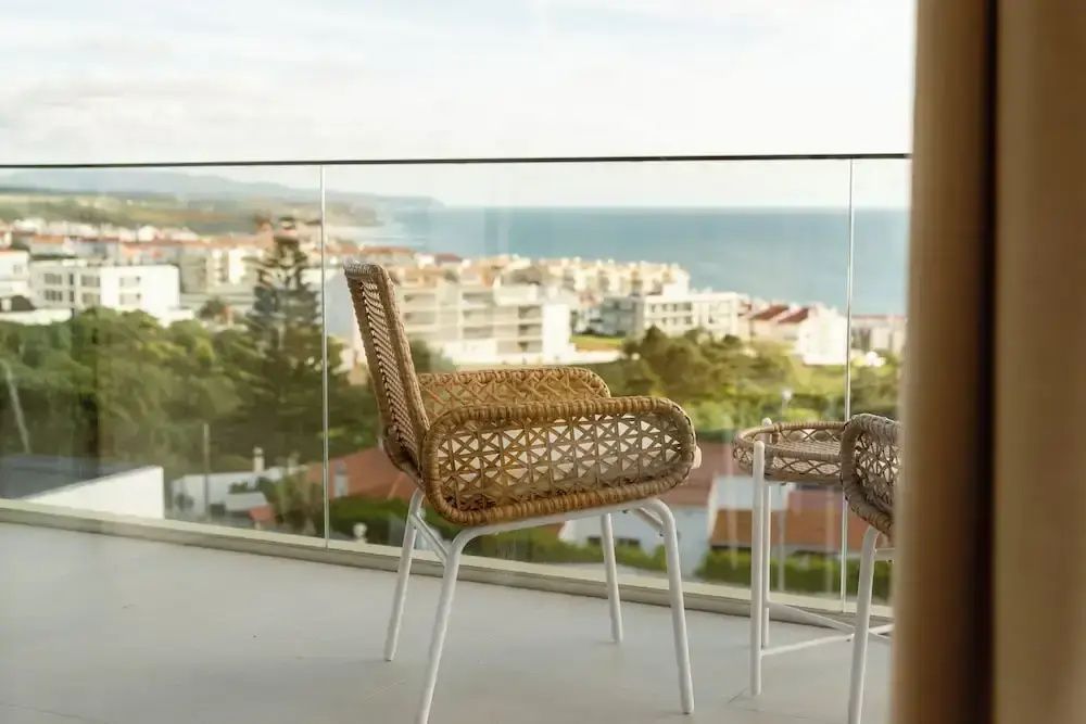 Balkon mit Flechtstuhl, Beistelltisch, Glasgeländer und Aussicht auf das Meer.