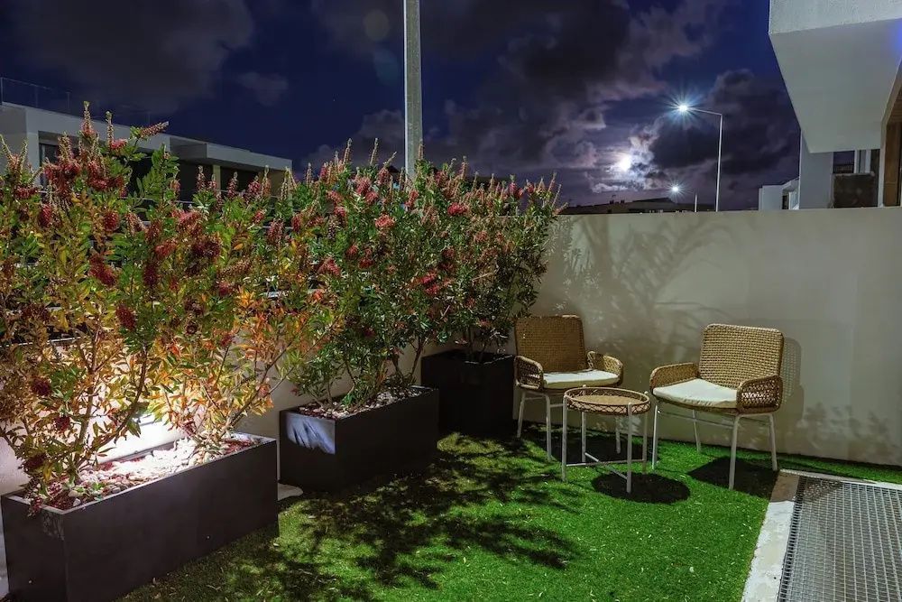 Beleuchtete Terrasse mit Kunstrasen, zwei Flechtsesseln und Blühpflanzen im Kübel bei Nacht.