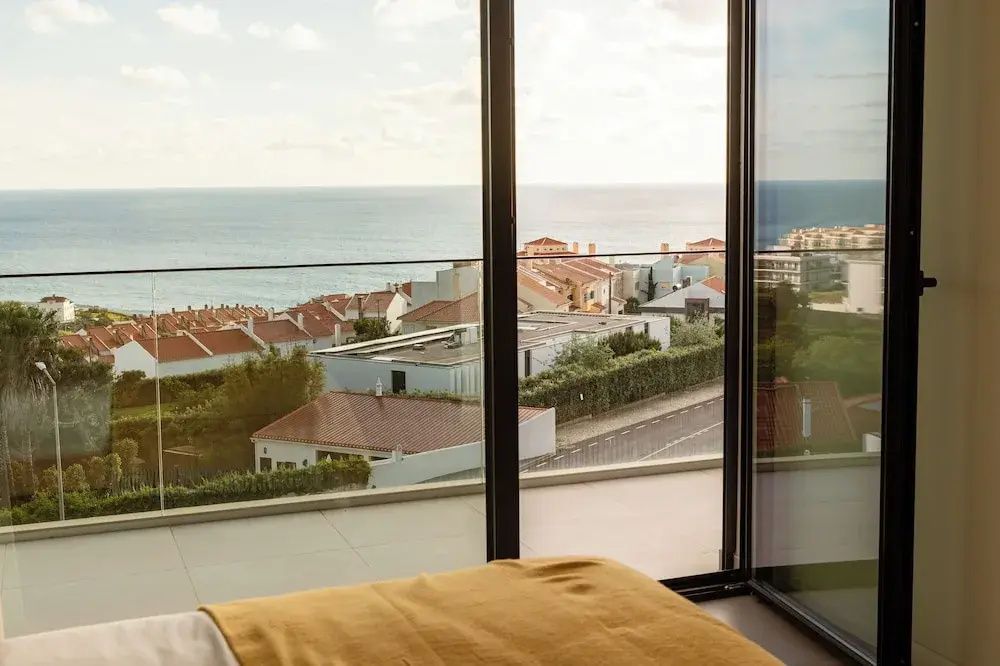 Blick durch ein großes Fenster auf Küstenhäuser und das ruhige Meer dahinter.