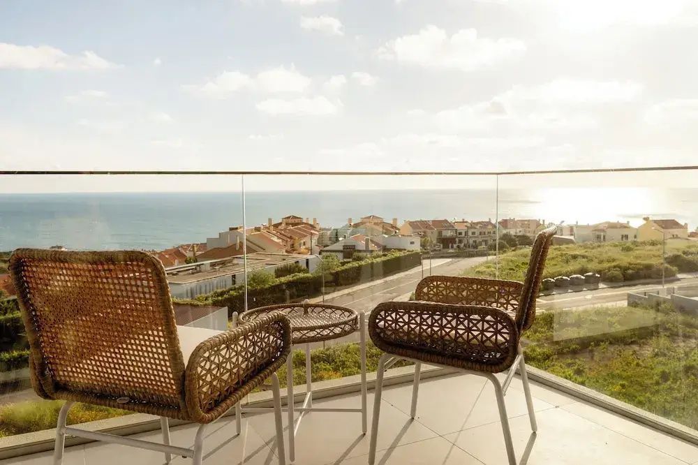 Balkon mit zwei Flechtstühlen, Glastisch und Glasgeländer mit Meeresblick.