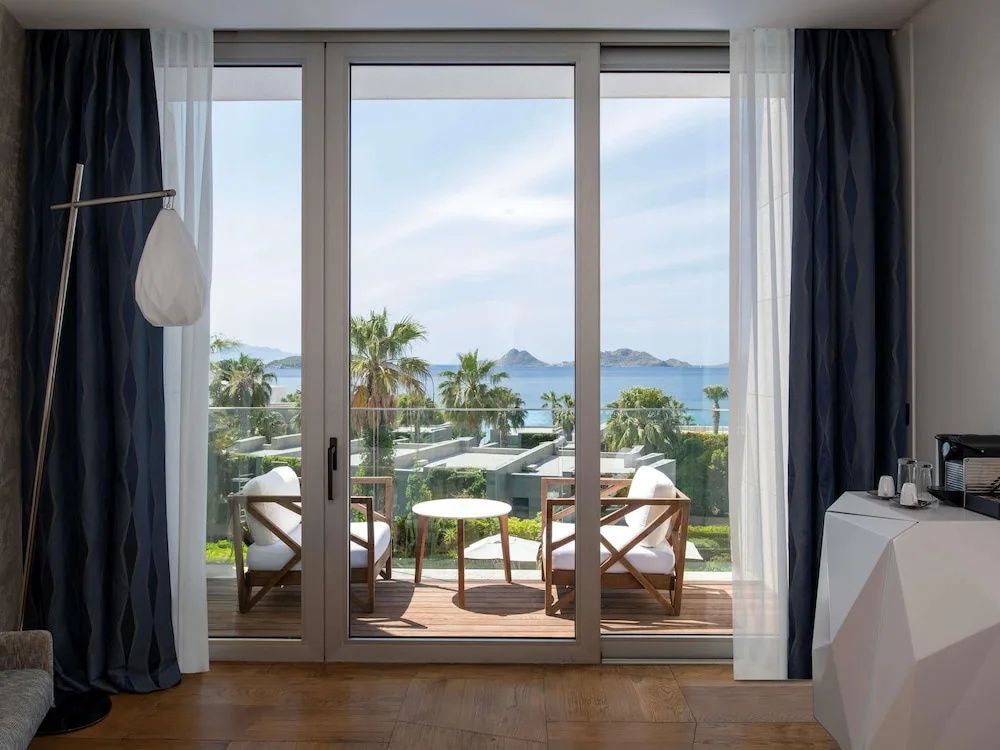 Blick durch ein Balkonfenster auf eine möblierte Terrasse mit Ausblick auf Palmen und das Meer.