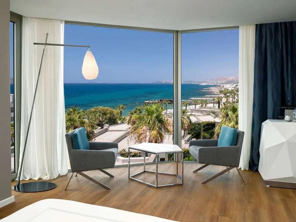 Sitzbereich mit zwei Sesseln vor großen Fenstern mit Panoramablick auf das blaue Meer.