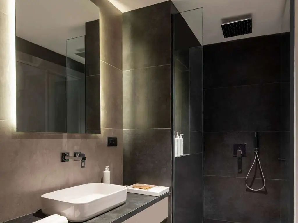 Modernes Badezimmer mit beleuchtetem Spiegel, weißem Waschbecken und dunkler Duschkabine.