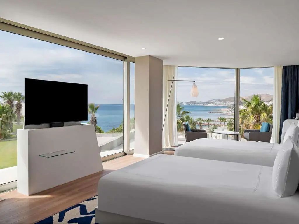 Helles Hotelzimmer mit zwei Betten, Fernseher und großer Fensterfront mit Blick auf das Meer.