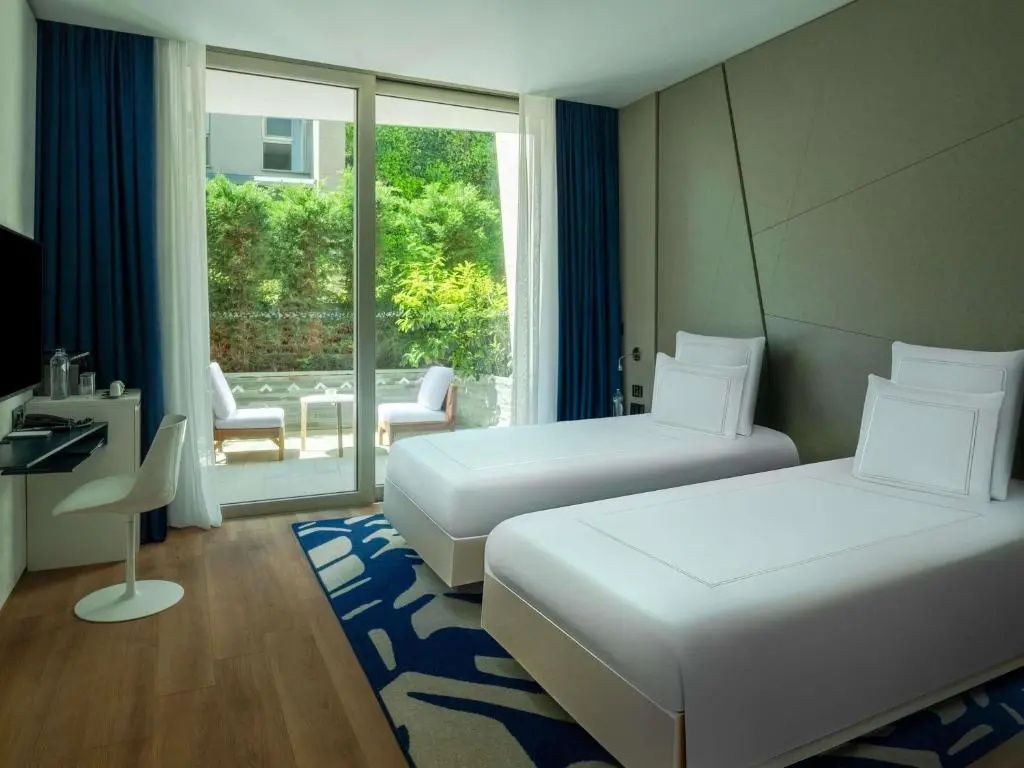Zimmer mit zwei Einzelbetten, blauem Teppich und Balkon mit Gartenblick.