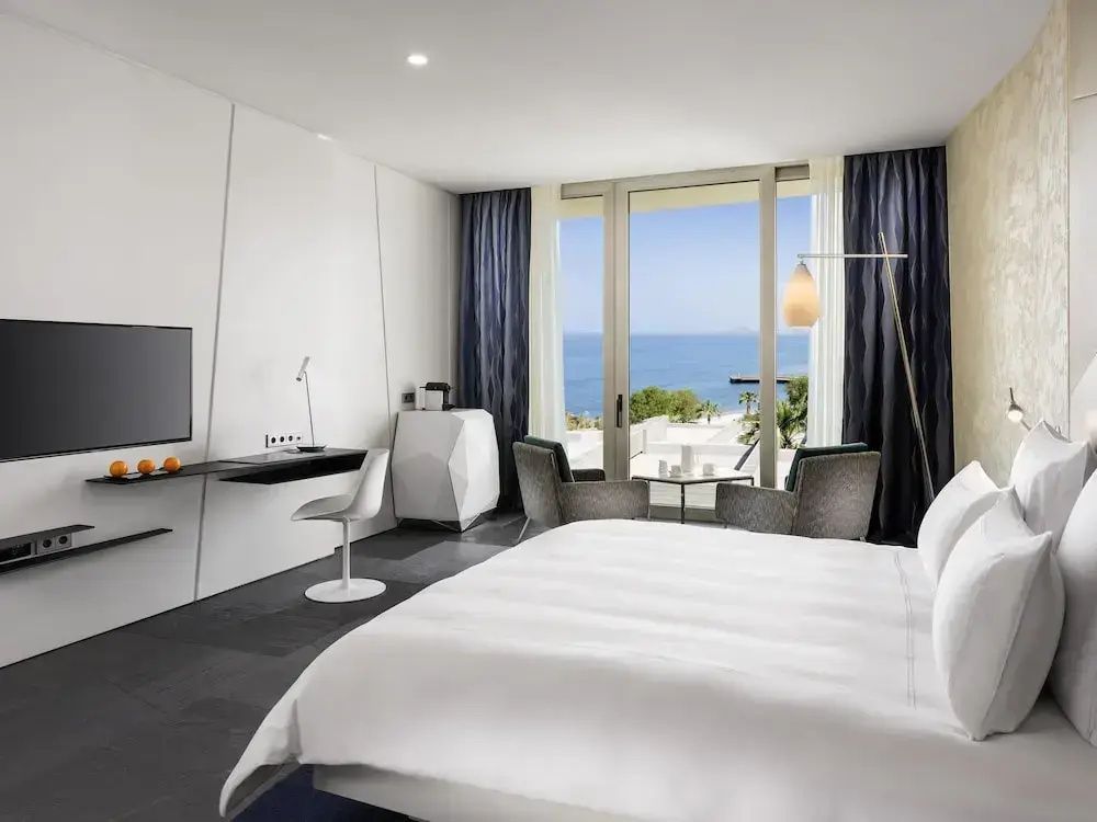 Zimmer mit hellweißem Doppelbett, Schreibtisch und freiem Blick auf das blaue Meer.