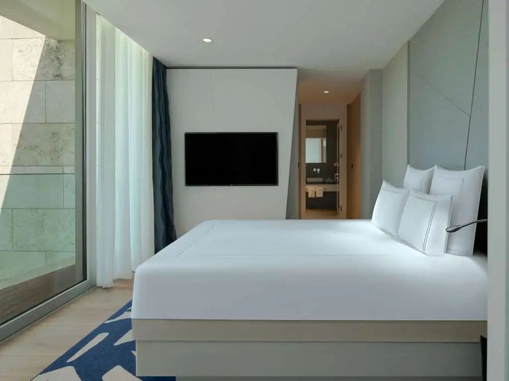 Schlafbereich mit weißem Doppelbett, Wand TV und Durchgang zum Badezimmer.