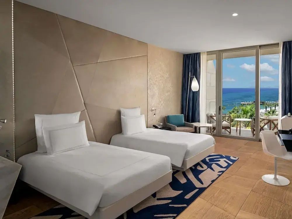 Modernes Hotelzimmer mit zwei Einzelbetten, blauem Teppich und Zugang zum Balkon mit Meerblick.