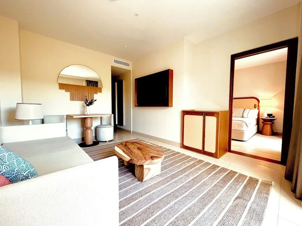 Wohnbereich einer Suite mit Sofa, Holztisch, Fernseher an der Wand und Blick ins Schlafzimmer.