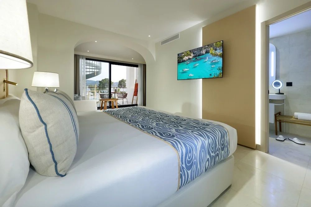 Helles Hotelzimmer mit Doppelbett, Wandfernseher und Blick auf den Balkon.