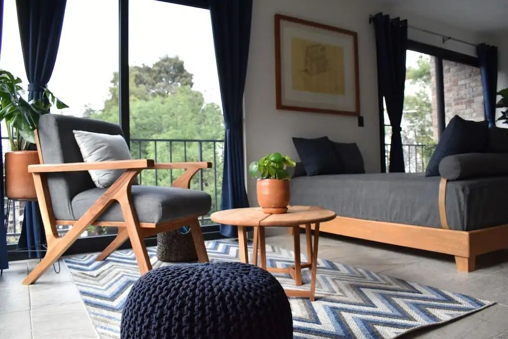 Modernes Wohnzimmer mit Sessel, Holztisch, dunkelblauem Pouf und Teppich vor großem Fenster.