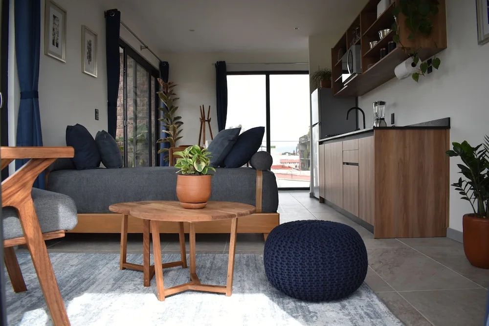 Moderne Sitzecke mit Holzbeistelltischen, grauem Sofa, blauem Pouf und integrierter Küchenzeile.
