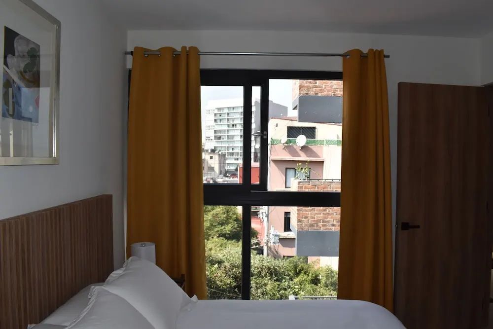 Bett im Hotelzimmer mit gelben Vorhängen und Blick aus dem Fenster auf Stadtgebäude.