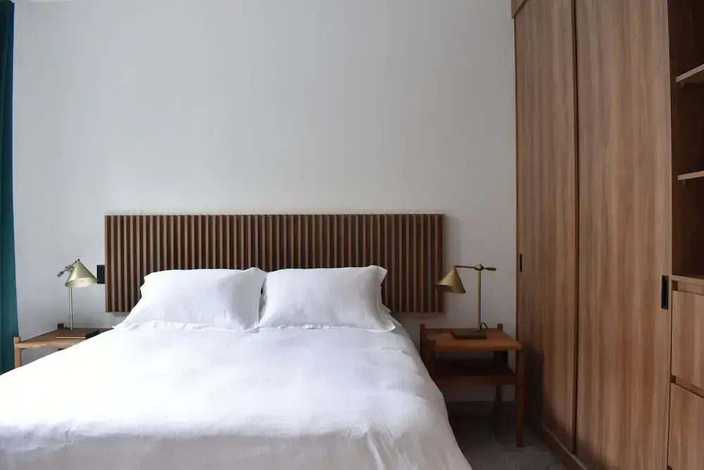 Doppelbett mit weißer Bettwäsche, Holzkopfteil, Nachttisch und Holzschrank im Hotelzimmer.