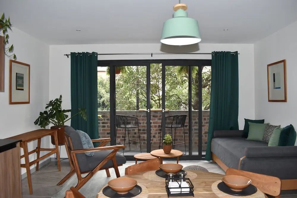 Wohnbereich mit Holztisch, Sessel, grünen Vorhängen und großer Glasfront zum Balkon.