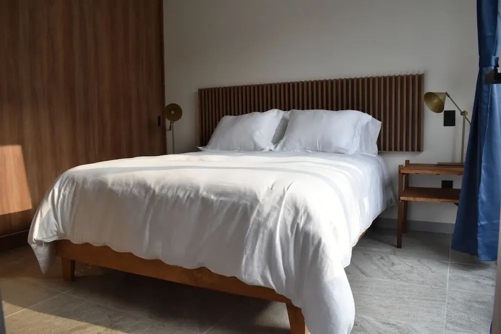 Doppelbett mit weißem Bezug, Holzrahmen und hölzernem Kopfteil auf hellem Fliesenboden.