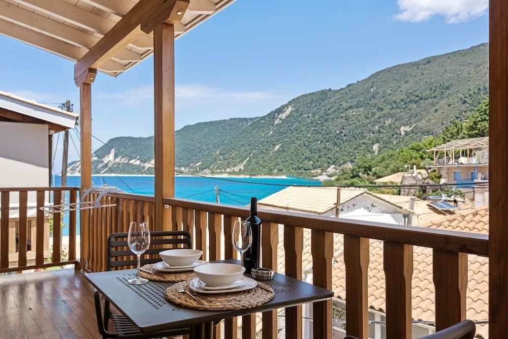 Tisch auf einem Holzbalkon mit zwei Gläsern und Blick auf das blaue Meer und grüne Berge.