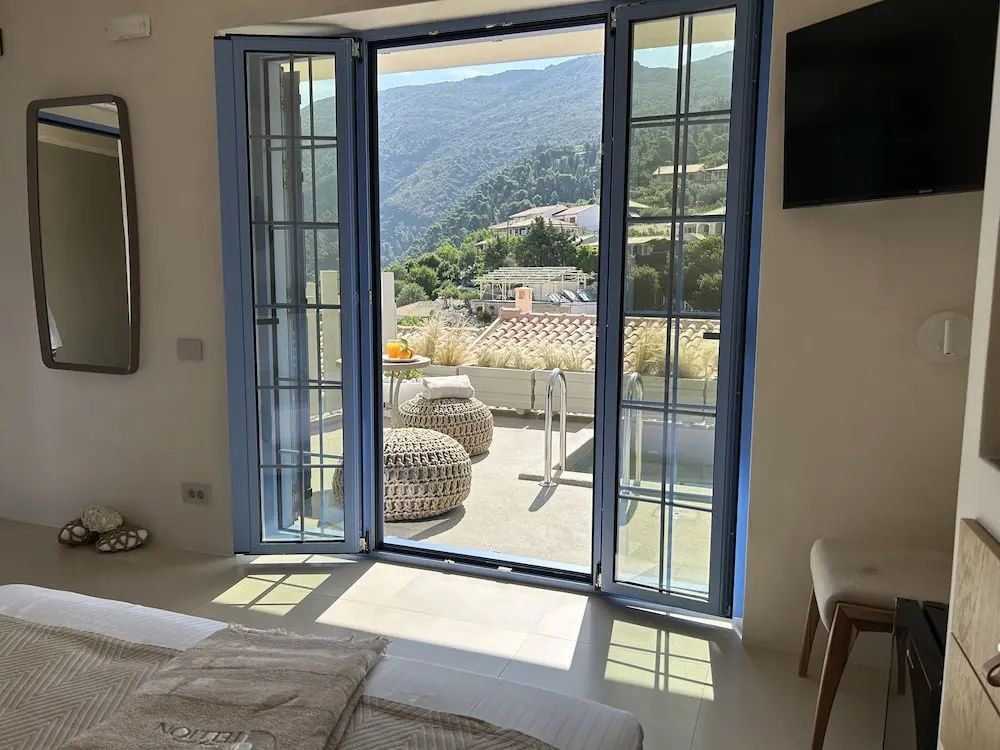 Offene blaue Balkontür mit Blick auf Terrasse, kleinen Pool und bewaldete Berge im Hintergrund.