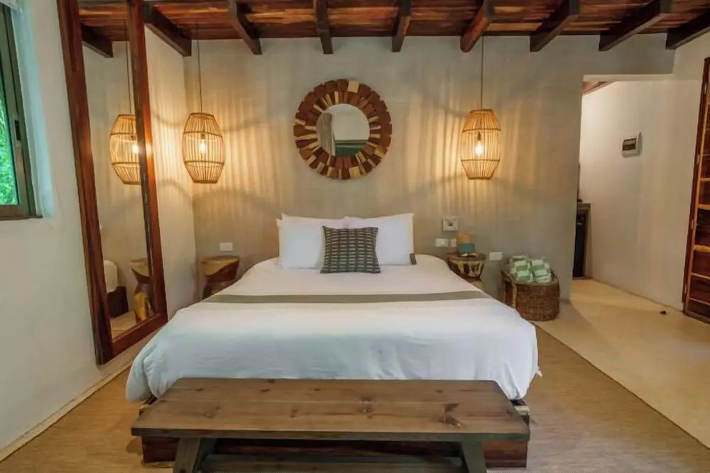 Doppelbett mit weißer Bettwäsche, Holzbank, Rattanleuchten und rundem Wandspiegel.