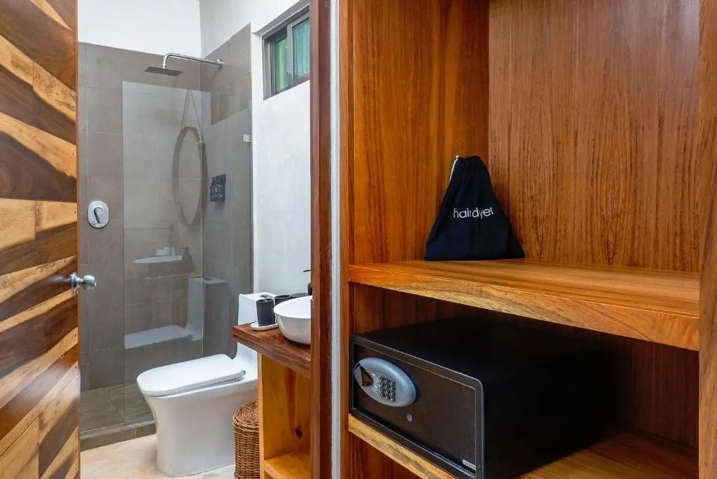 Ein geöffneter Holzschrank mit Zimmersafe vor einem Badezimmer mit Dusche und WC.