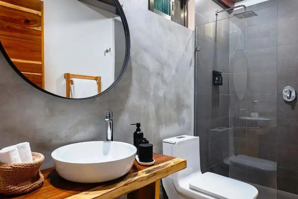Badezimmer mit weißem Aufsatzwaschbecken auf Holzplatte, rundem Spiegel und Glasdusche.