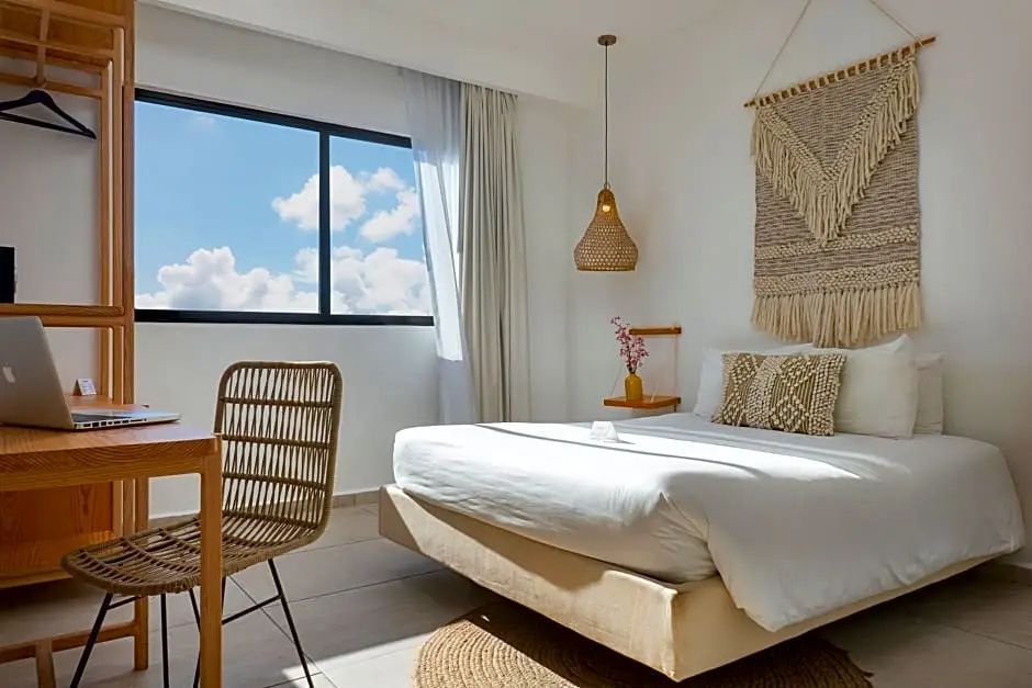 Hotelzimmer mit hellen Möbeln, Stuhl, Flechtlampe und Fenster zum Himmel.