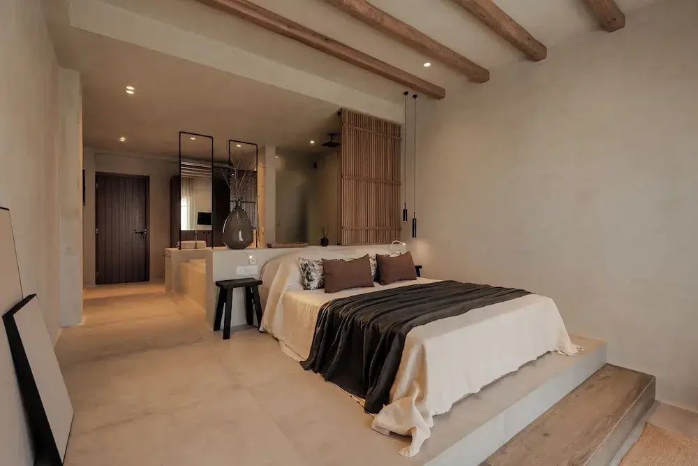 Doppelbett in hellem Hotelzimmer mit Holzbalken an der Decke und offenem Badbereich dahinter.