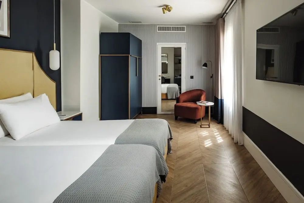 Hotelzimmer mit zwei Betten, Parkettboden, rotem Sessel am Fenster und blauem Kleiderschrank.