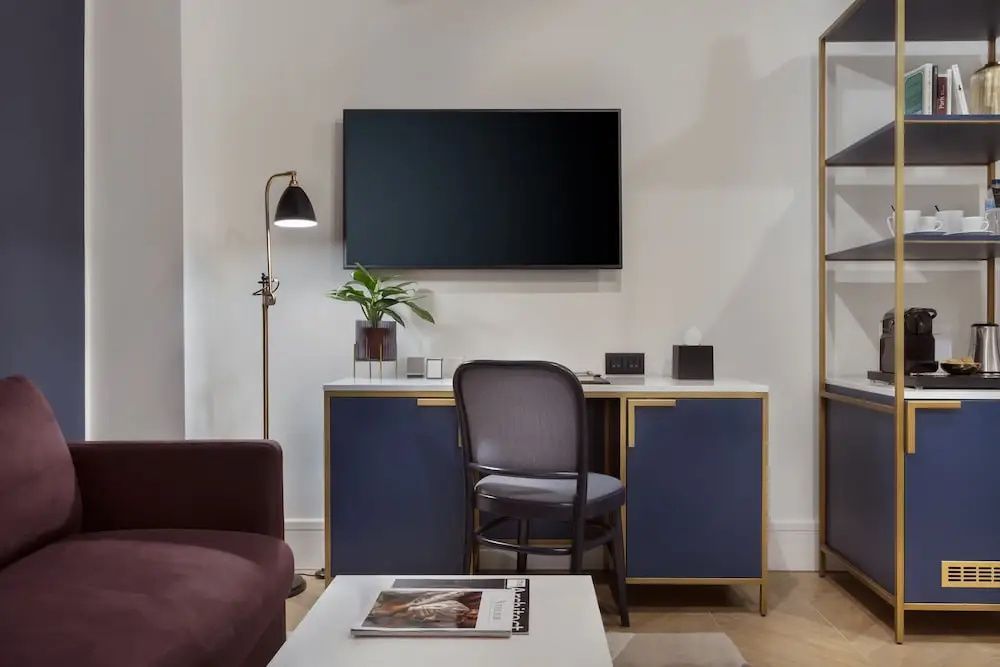 Arbeitsbereich im Hotelzimmer mit blauem Sideboard, Stuhl, Wandfernseher und Stehlampe.