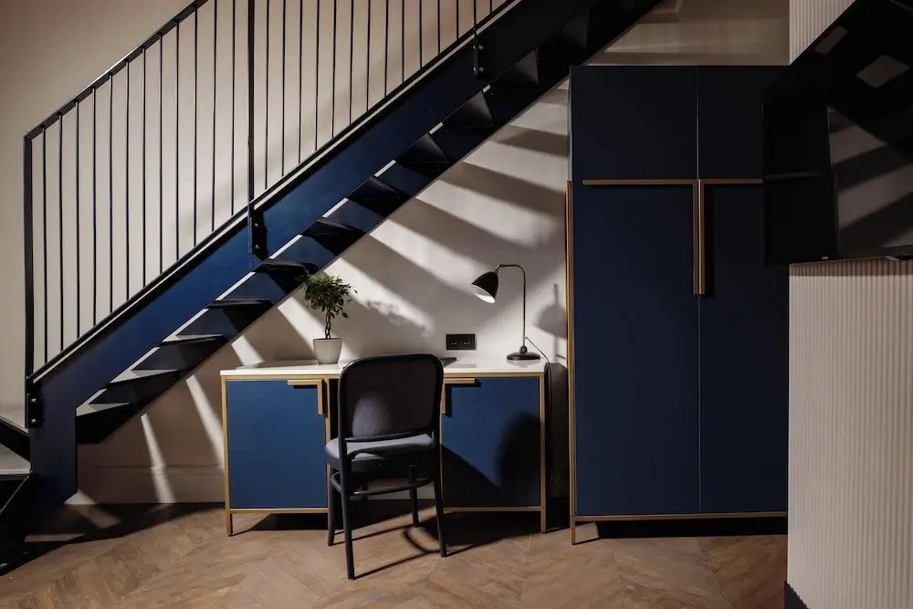 Schreibtischbereich mit Stuhl und Lampe unter einer dunklen Treppe im Hotelzimmer.