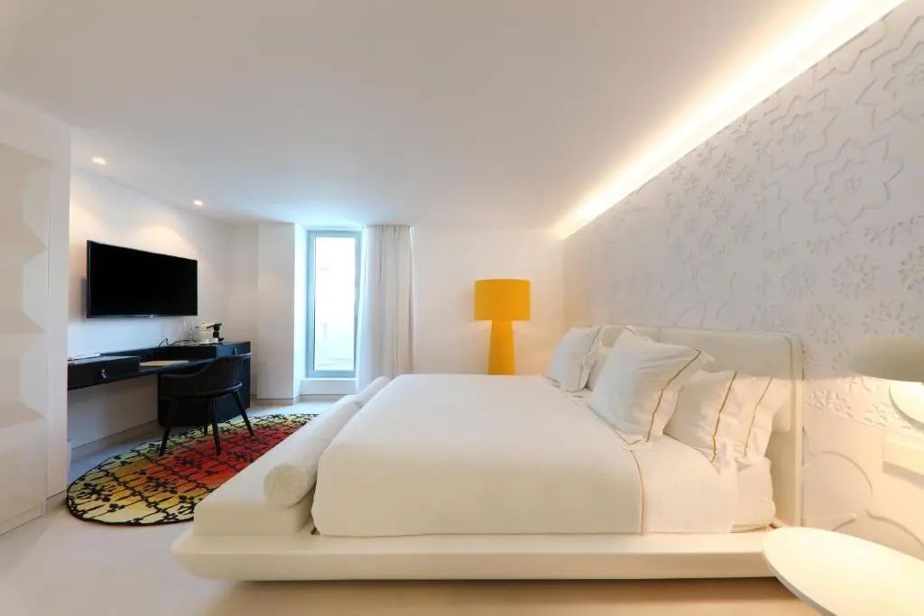Helles Hotelzimmer mit weißem Doppelbett, gelber Stehleuchte und buntem Teppich
