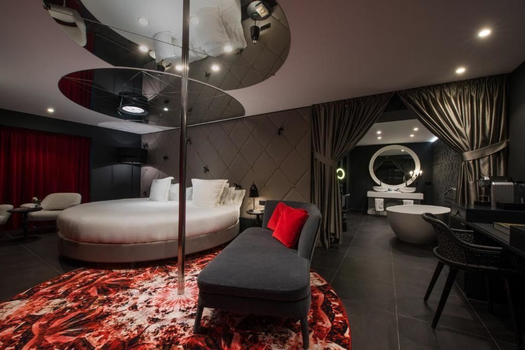 Rundes Bett unter einem Decken-Spiegel, grau-rotes Liegesofa und eine Tanzstange im dunklen Zimmer.