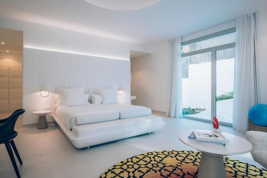 Hell gestaltetes Schlafzimmer mit weißem Doppelbett, runden Tischen und Glasfront.
