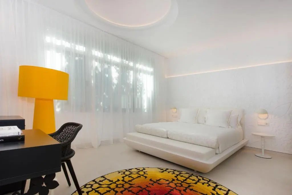 Weiß eingerichtetes Hotelzimmer mit einem großen Bett, gelber Lampe und buntem Teppich