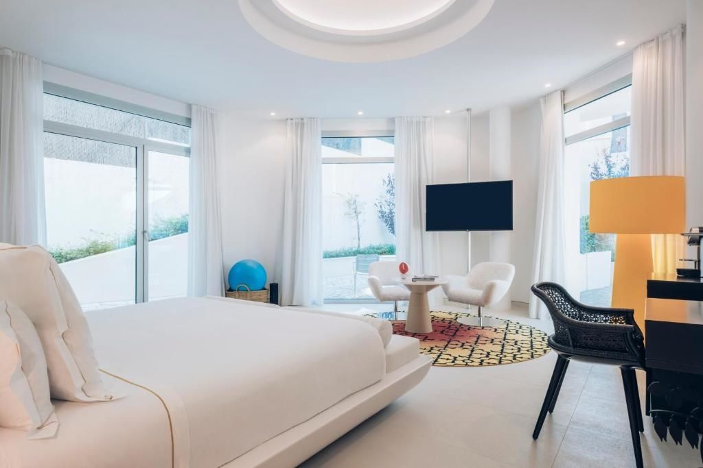 Geräumiges Zimmer mit Bett, Sitzecke, TV Bildschirm und großen Fensterfronten.