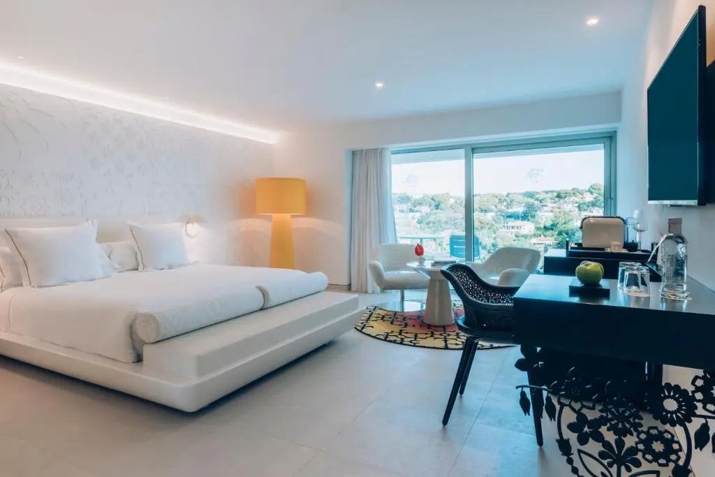 Weiß eingerichtetes Hotelzimmer mit Doppelbett, gelber Stehlampe und Fenster zur Terrasse.