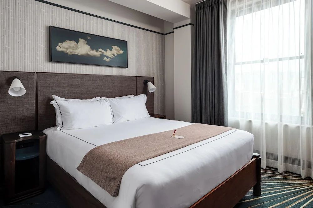 Hotelzimmer mit aufgemachtem Doppelbett, Vorhängen und einem Bild an der Wand.