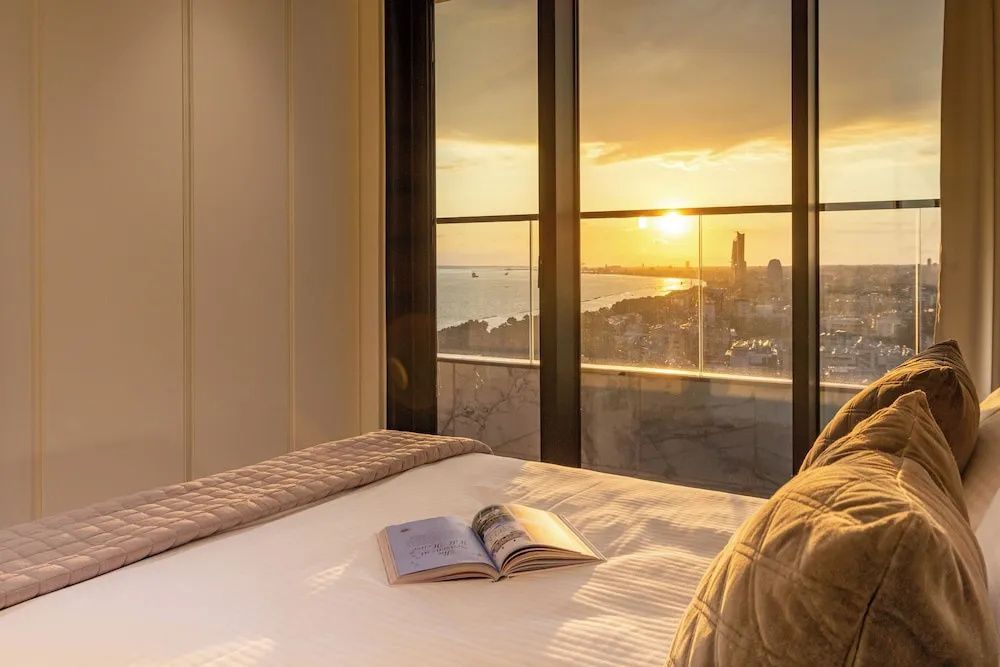Blick vom Bett durch eine große Fensterfront auf das Meer und die Küste beim Sonnenuntergang.