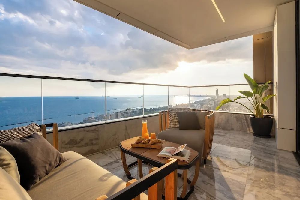 Balkon mit hölzernen Sesseln und Glasbrüstung mit Aussicht auf das Meer bei Sonnenuntergang.