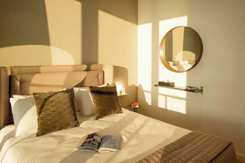 Helles Schlafzimmer mit Doppelbett, Kissen, Sonnenlicht an der Wand und rundem Spiegel.
