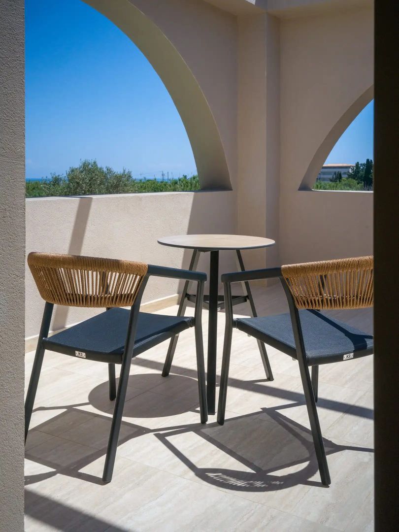 Balkon mit zwei Stühlen und einem runden Tisch unter Rundbögen mit Blick ins Grüne und aufs Meer.