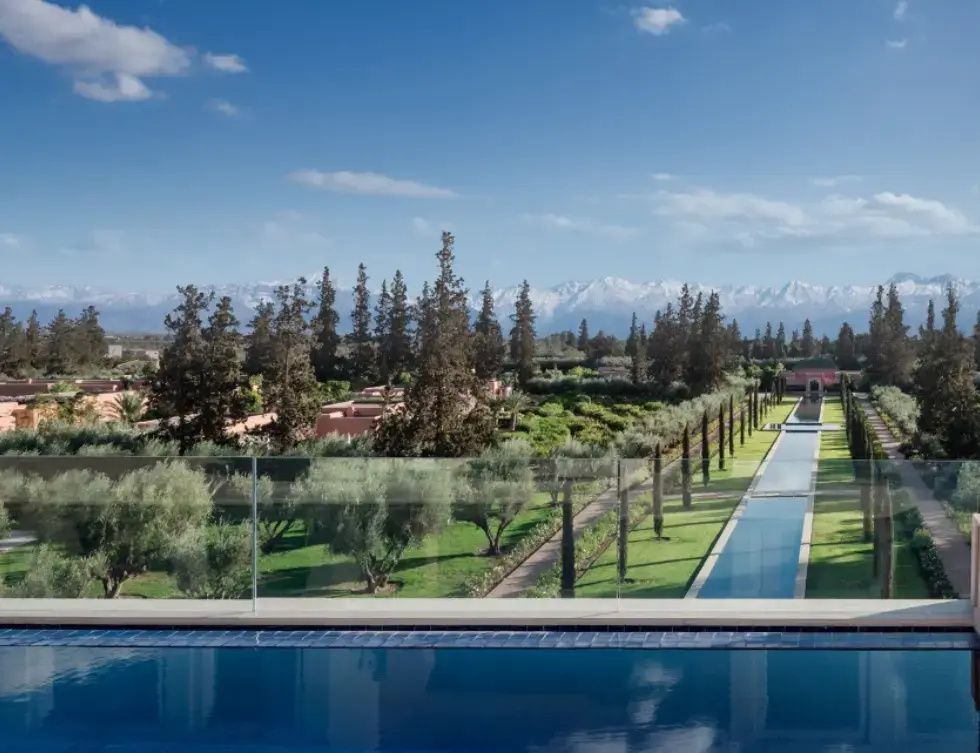 Blick über eine weitläufige Gartenanlage mit Wasserbecken auf verschneite Berge unter blauem Himmel.