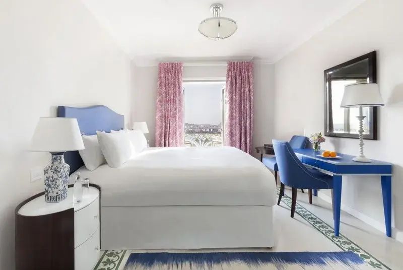 Helles Hotelzimmer mit Doppelbett, blauer Einrichtung, rosa Vorhängen und Balkonaussicht.