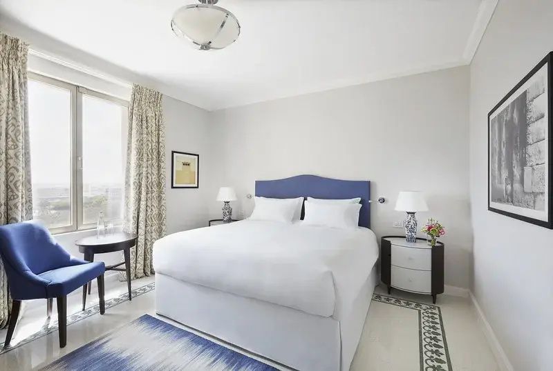 Helles Hotelzimmer mit weiß gedecktem Doppelbett, blauem Kopfteil und zwei Nachttischen.