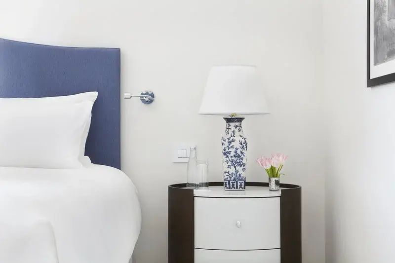 Runder Nachttisch neben dem Bett mit einer blau-weißen Porzellanlampe und rosa Blumen.